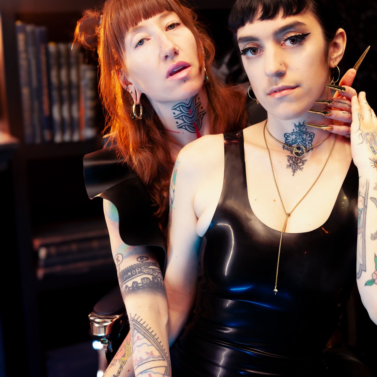 Two individuals with tattoos sitting together in a dimly lit room, wearing a gold o-ring choker and gold lariat necklace.