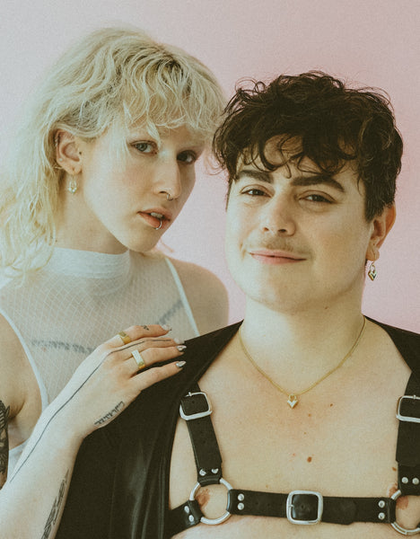 Two models wearing gold heart charm earrings, heart charm necklace and a harness