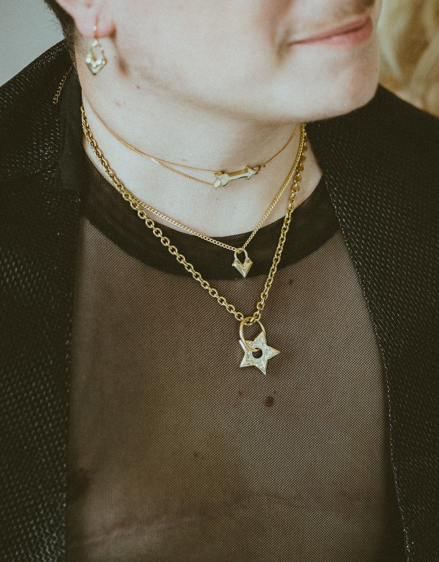 Close-up of model wearing black mesh shirt, gold arrow choker, heart charm choker and spur charm choker