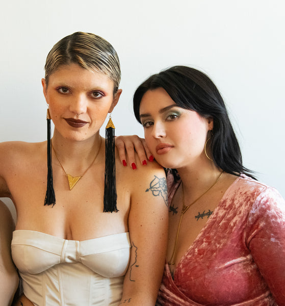 Model wearing starburst tassel earrings with matching choker, sitting beside another model in a pink velvet shirt and bolo tie