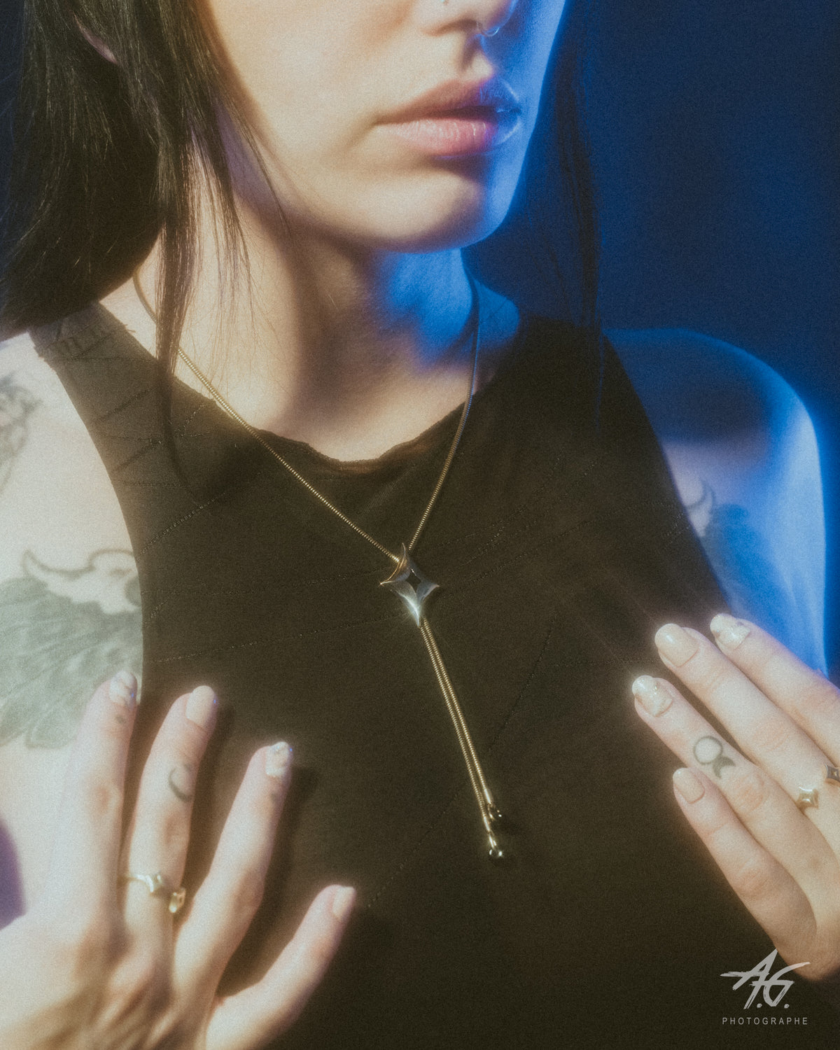 A close-up lifestyle shot of a model wearing a gold bolo necklace with a geometric star pendant, with a black sleeveless top under dramatic blue lighting.