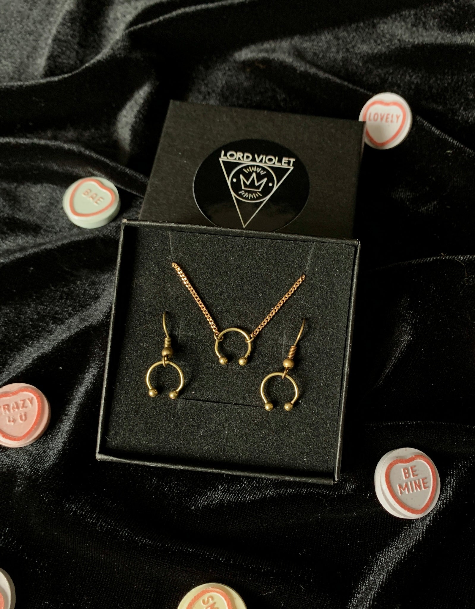 Gold barbell choker and earrings in a black gift box, surrounded by candy hearts on a black velvet background