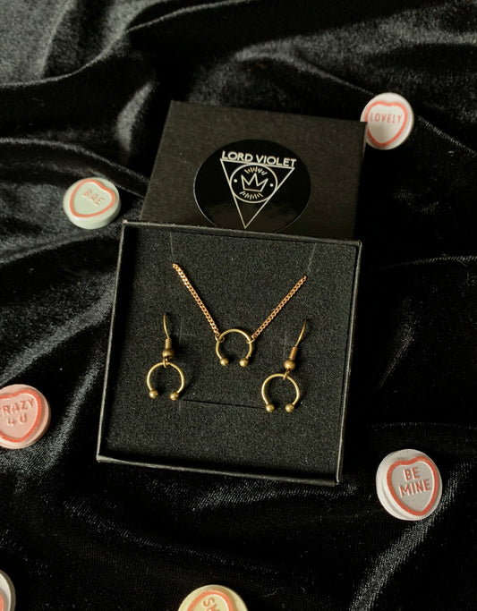 Gold barbell choker and earrings in a black gift box, surrounded by candy hearts on a black velvet background