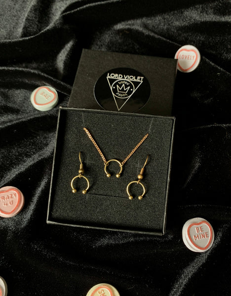 Gold barbell choker and earrings in a black gift box, surrounded by candy hearts on a black velvet background