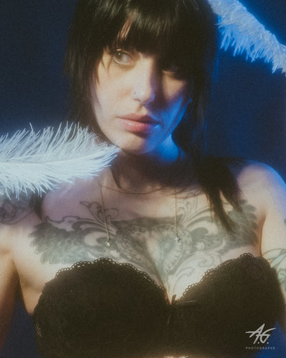 Close-up of a model wearing a delicate gold star charm choker a black lace bra, holding white feathers on each side of her face, against a dark blue background.