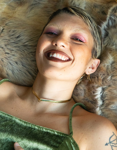 Model wearing gold cuff choker and green velvet dress, laying on beige fur rug