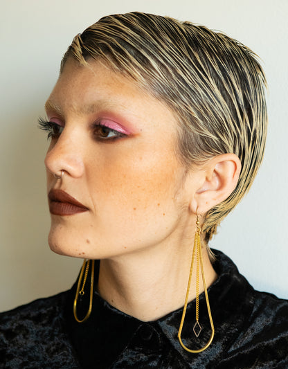 Side view of model wearing long arch earrings with triangle charms and black velvet button up shirt