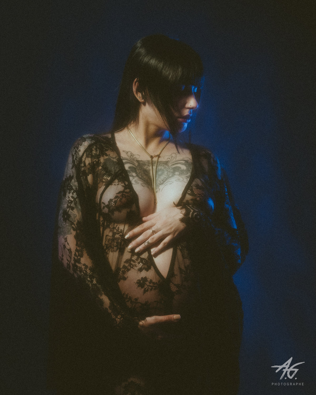 A pregnant model wearing a sheer black lace gown and a star bolo tie, against a dark blue studio background.