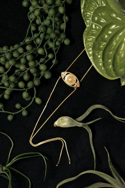 Gold bolo tie necklace with flower pendant on a black background, surrounded by plants