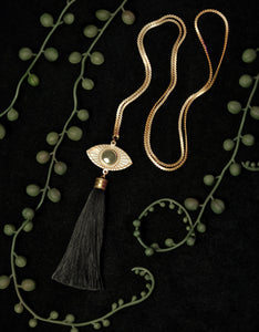 Gold eye pendant necklace with black tassel on a black background, surrounded by string of pearl plant