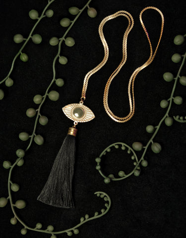Gold eye pendant necklace with black tassel on a black background, surrounded by string of pearl plant