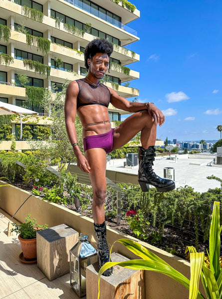 Model on a rooftop wearing gold belly chain, black mesh crop top, black boots and purple swim shorts