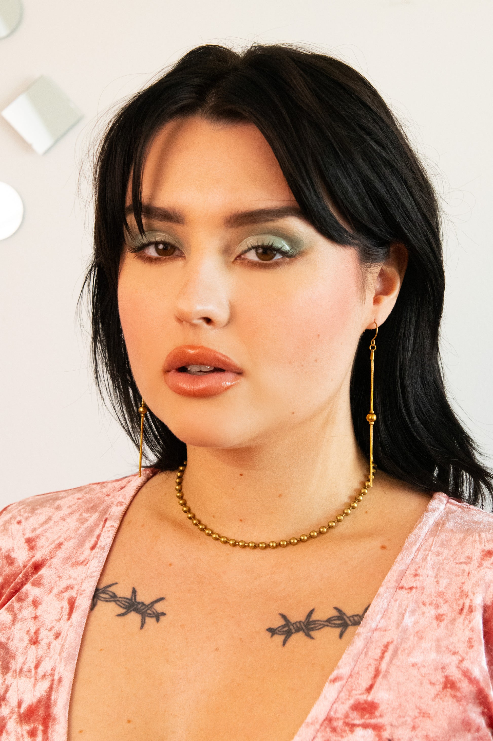 Model wearing shiny gold drop earrings, ball chain choker and pink velvet top