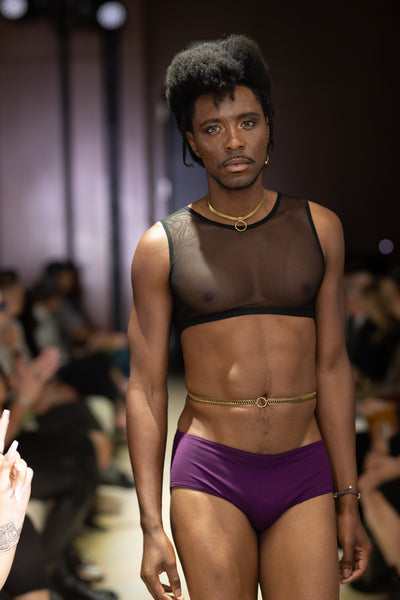 Model on runway wearing gold belly chain, black mesh crop top , gold choker and purple swim shorts