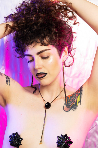 Model wearing gold tassel earrings, bolo tie, and black rhinestone nipple pasties while holding up her hair